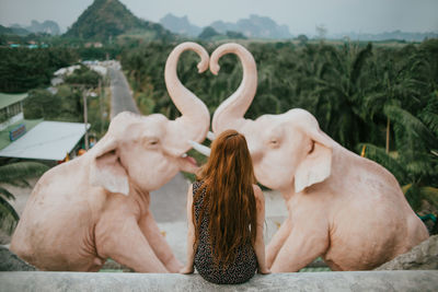 Back view of anonymous female traveler sitting near statue of elephants against lush green trees and mountains in tropical country