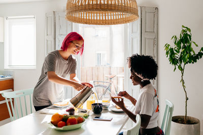 Young woman with dyed hair smiling and filling mug with fresh coffee while having breakfast in morning at home