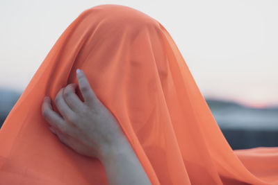 Close-up of woman covering face with orange fabric against sky