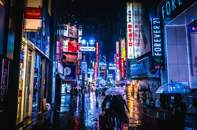 People on street amidst illuminated buildings during rainy season at shibuya