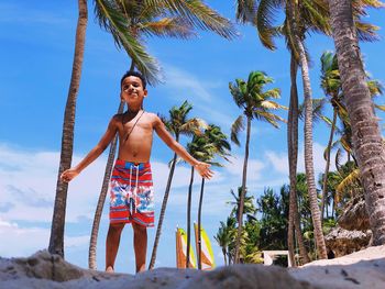 Portrait of shirtless boy standing at beach against palm trees