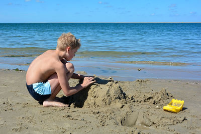 Little blond boy with swimming trunks builds a sand castle in the sand on the beach 