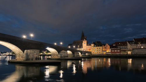 Bridge over river by illuminated buildings against sky at night