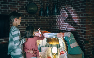 Two children in pajama having fun playing with shadow puppets over brick wall at home