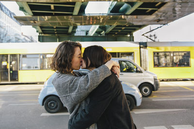 Lesbian couple kissing while standing on street under bridge in city