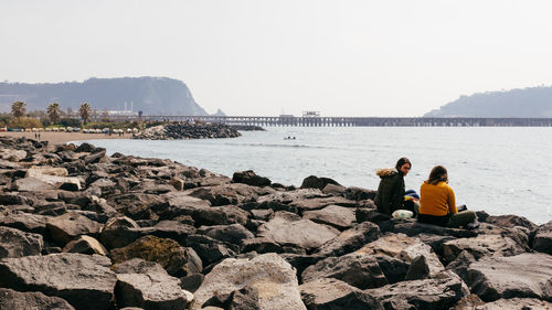 Rear view of people sitting on rock by sea against sky