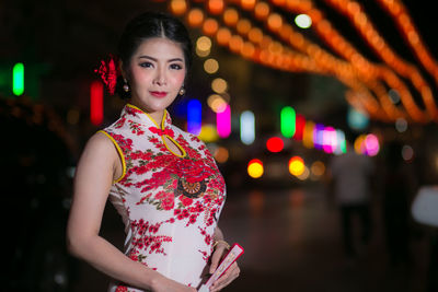 Portrait of young woman standing against illuminated lights at night