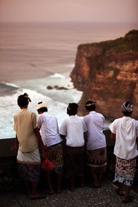 Rear view of boys wearing headdresses standing by retaining wall against sea during sunset