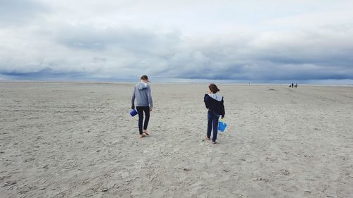 Rear view of friends with buckets walking on beach against cloudy sky