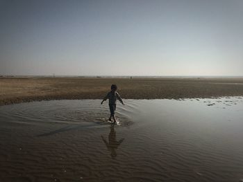 Boy walking in shallow water at beach against clear sky