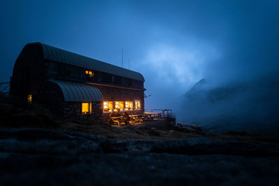Group of unrecognizable adventurers sitting on terrace of guest house with illuminated windows located in mountainous area in foggy night