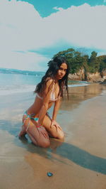 Portrait of young woman in bikini kneeling on beach against sky