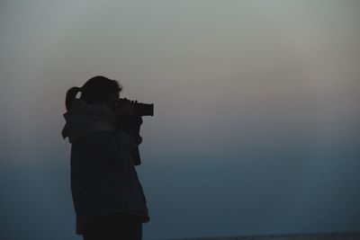 Side view of woman photographing with camera against sky during foggy sunset