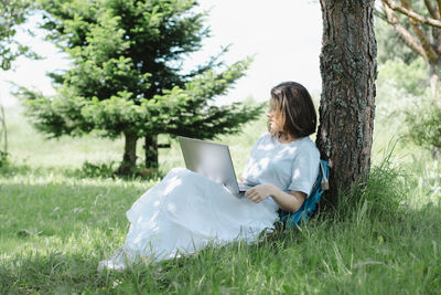 Nice teenage girl siting near tree in summer park with open laptop in her hands and studying