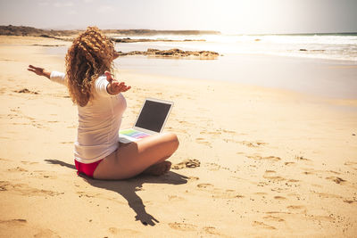 Full length of seductive woman with arms outstretched using laptop at beach