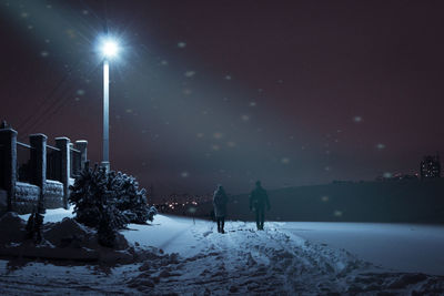 Two people are walking on a snowy road at night by the light of lanterns