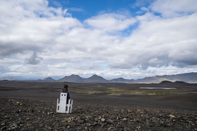 Rear view of woman sitting on chair at landscape against cloudy sky