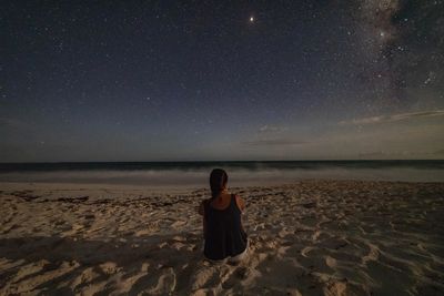 Rear view of man sitting on beach against sky at night