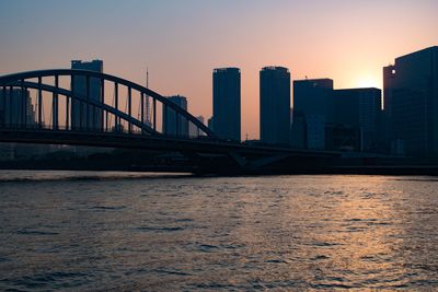 Bridge over river by buildings against sky during sunset