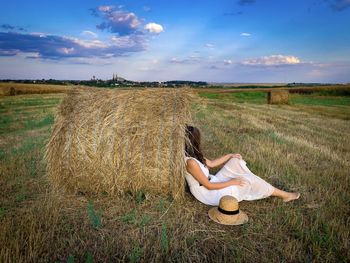 Rural scene of woman sitting down on the ground and leaning against a haystack on the field