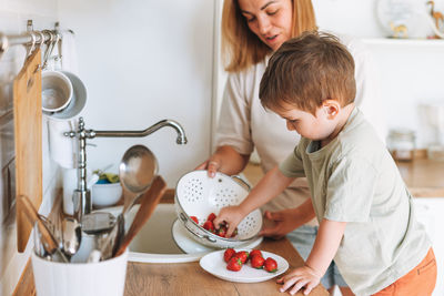 Young woman mother with her toddler boy son eat srawberries in kitchen at home