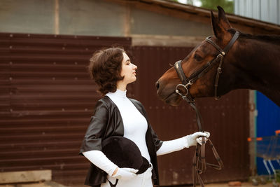 Young woman rider with brown beautiful horse, stands nearby in jockey clothes