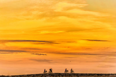 Cyclists riding on field against orange sky