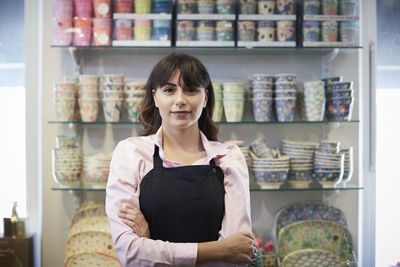 Portrait of confident saleswoman standing with arms crossed against crockery at boutique