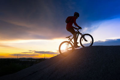 Man riding bicycle on street against sky during sunset