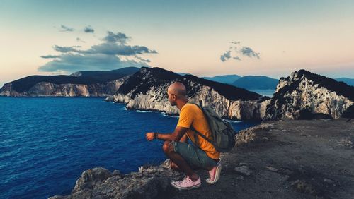 Side view of man looking at sea while crouching on mountain during sunset