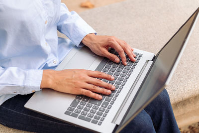 From above cropped unrecognizable female entrepreneur hands typing on laptop while sitting on stone bench in park and working remotely