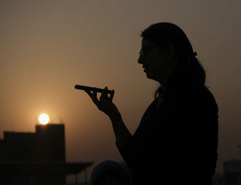 Side view of silhouette man holding camera against sky during sunset