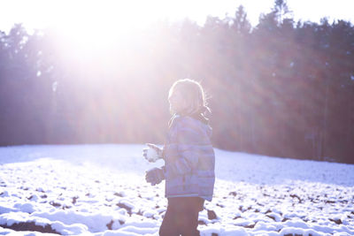 Side view of boy holding snowball while standing on field during sunset