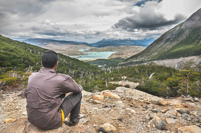 Rear view of man sitting on mountain against cloudy sky