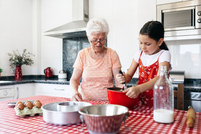 Granddaughter with whisk near grandmother in eyewear cooking dough at table in house