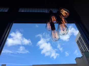 Low angle view of illuminated light bulbs hanging in building against sky