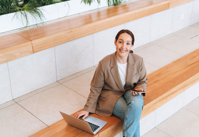 Adult smiling business woman in stylish beige suit and jeans working on laptop at public place