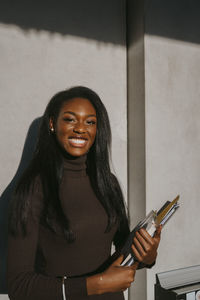 Portrait of cheerful young woman holding book standing against gray wall on sunny day