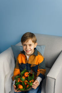 Boy cleans and eats a tangerine while sitting in a chair on a blue background.