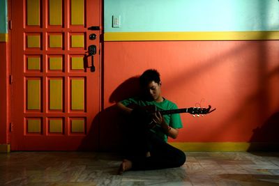 Young man playing guitar while sitting against wall