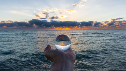 Cropped hand person holding crystal ball by sea against sky during sunset