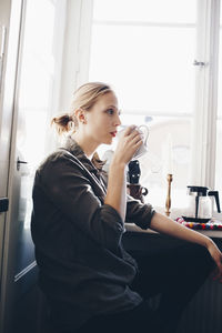 Side view of woman drinking coffee against window in kitchen at home