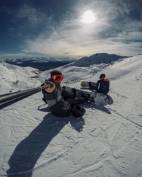 People with snowboards sitting on snowcapped mountain against sky