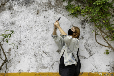 Woman wearing mask taking selfie standing against wall