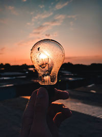 Close-up of hand holding light bulb light against sky during sunset