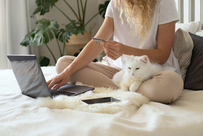 Midsection of paying online sitting on bed with cat