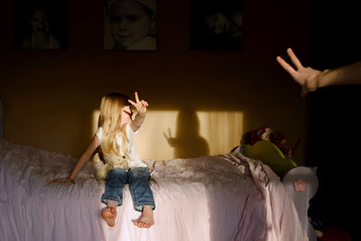 Happy girl showing peace sign while sitting on bed at home