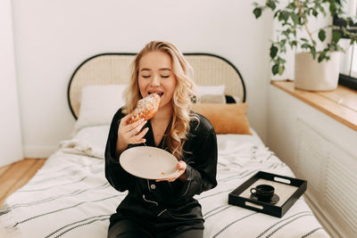 A happy pretty young woman in pajamas is resting and having breakfast in bed in a cozy apartment