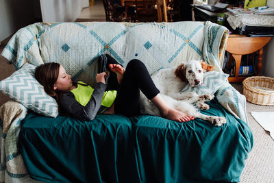 Side view of girl using digital tablet while lying with dog on sofa at home