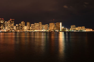 Illuminated buildings by sea against sky at night